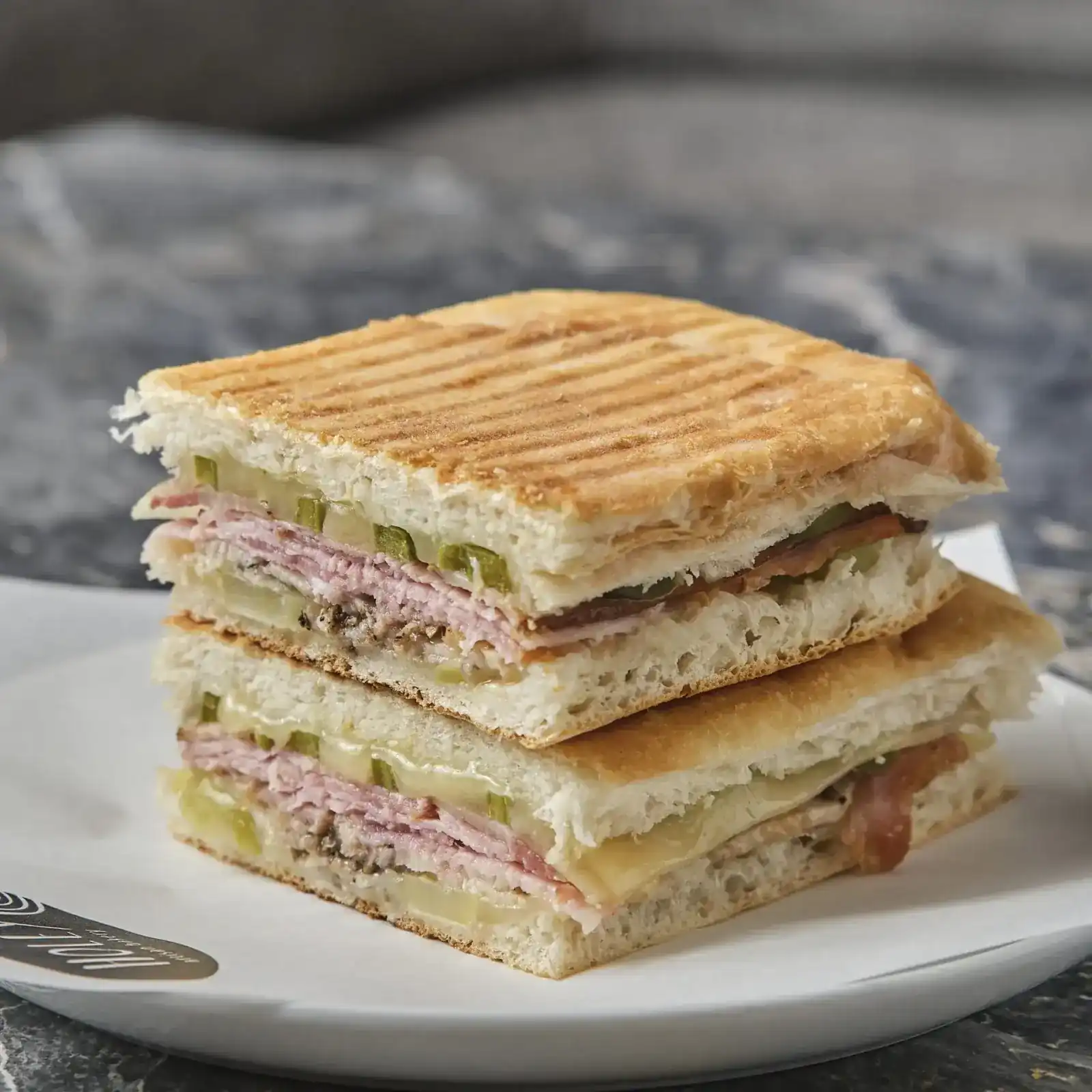A pressed Cubano sandwich cut in two and stacked on a white plate, on dark marble