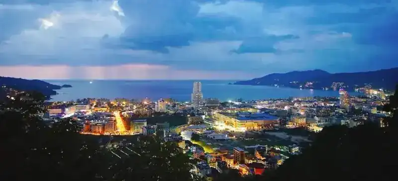 Patong Bay at dusk under storm clouds, the town lights coming on below the hill