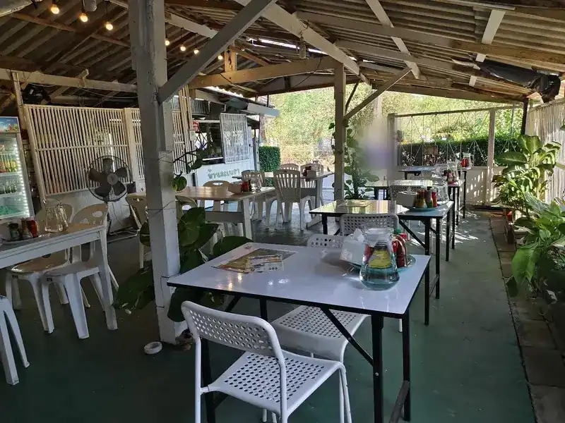 The open-air dining room: white tables and chairs, water jugs, fans and potted plants under a timber and tin roof