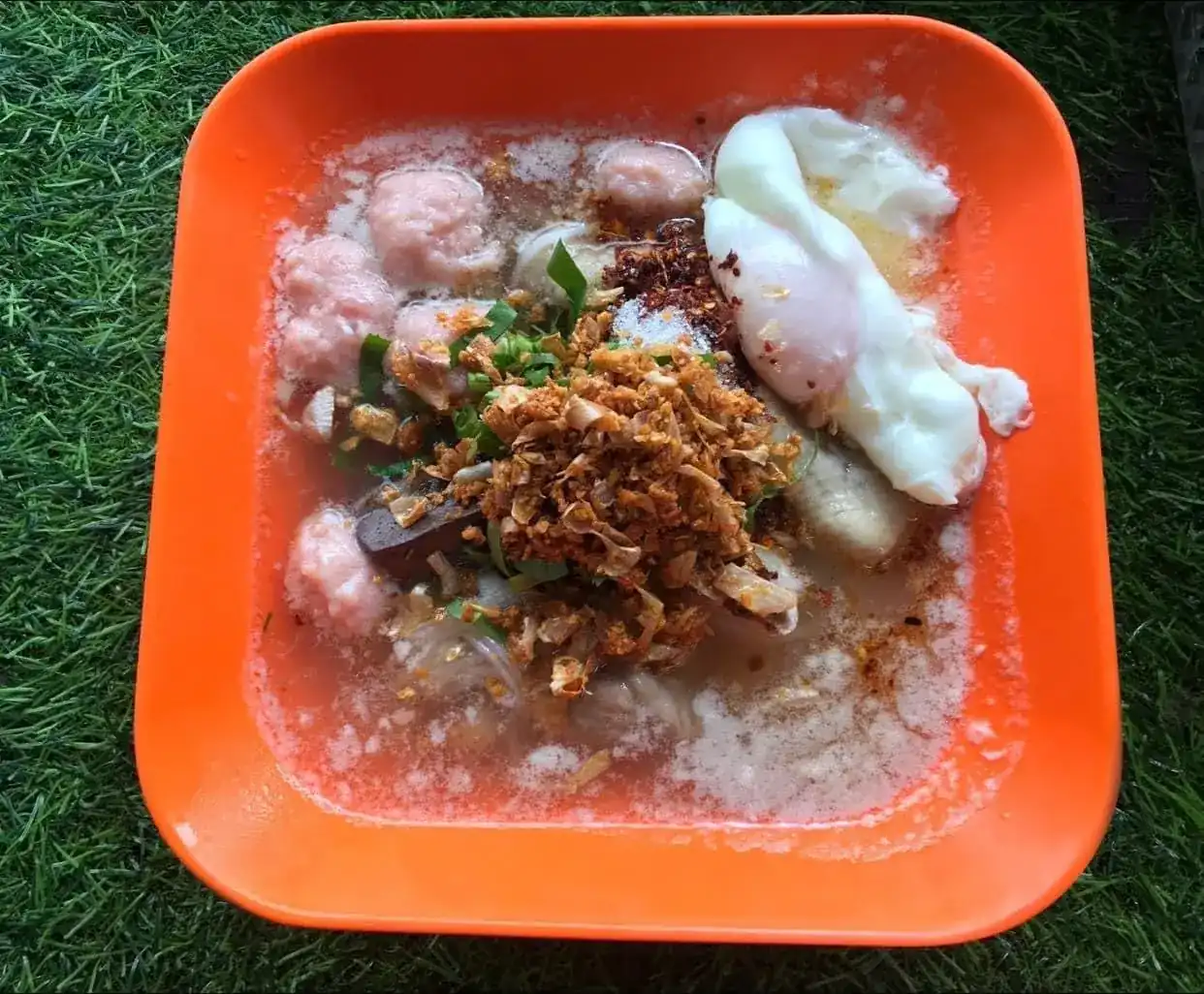 Kuay jap in an orange square bowl with pork balls, a soft-boiled egg, pork blood and a heap of fried garlic