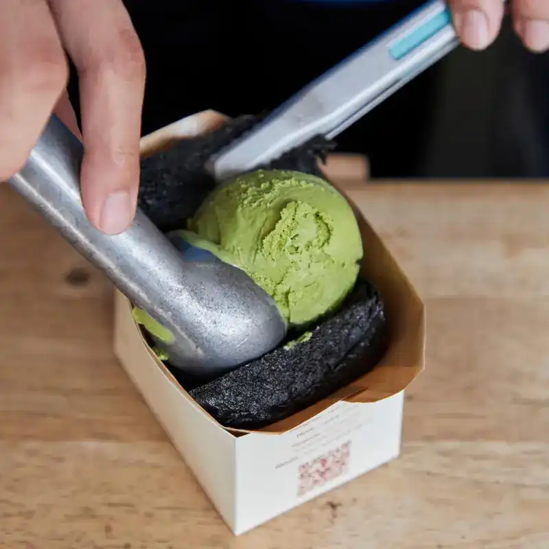 A scoop pressing matcha ice cream onto a black grilled bun in a paper tray