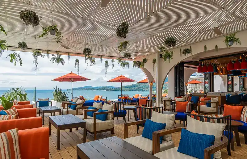 The open-air dining terrace with hanging plants, red parasols and the bay behind