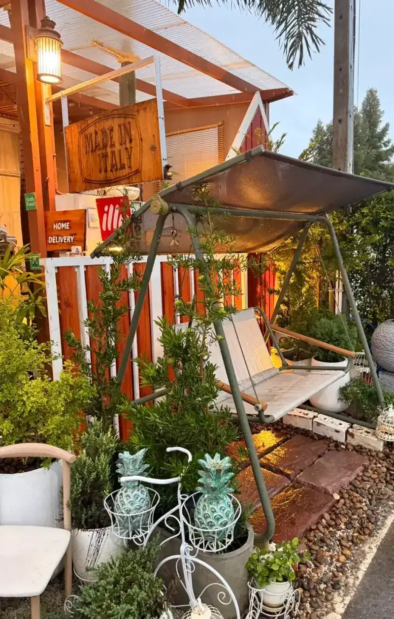 The garden front at dusk: the carved Made in Italy sign, a Home Delivery board, a garden swing and potted plants