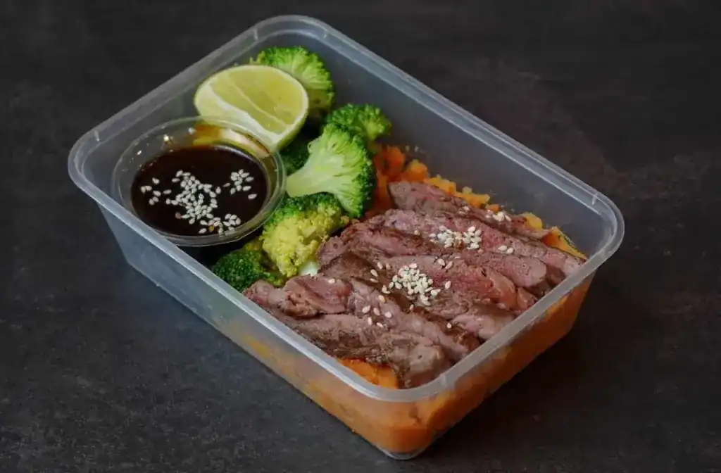 Sliced beef striploin with sesame, broccoli, lime and teriyaki sauce over sweet mash, in a clear meal-prep box