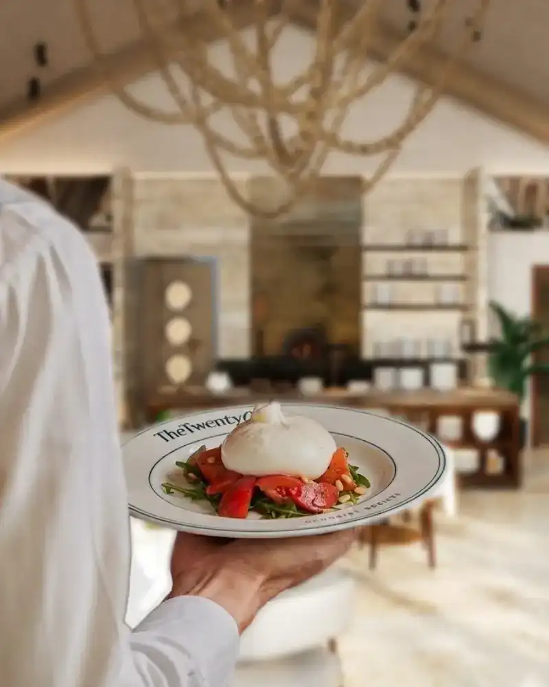 A plate of burrata and tomatoes carried through the dining room, the wood-fired oven glowing behind