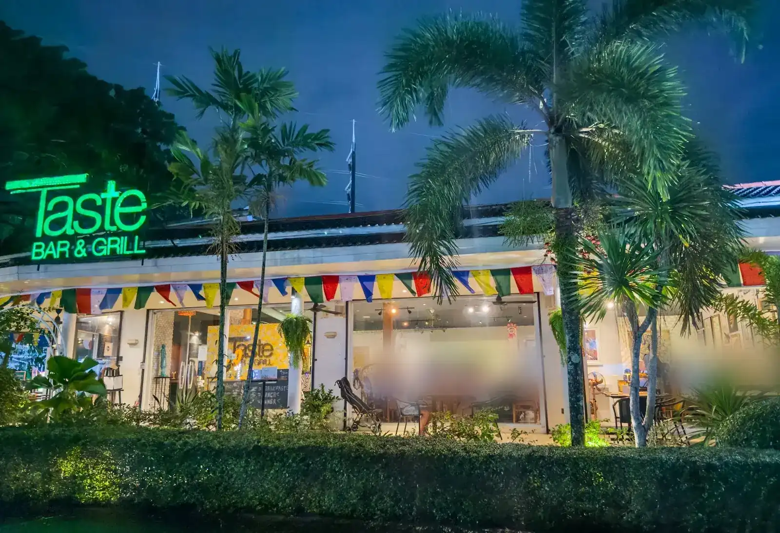 Taste's green neon sign above the open-fronted restaurant at night, with palm trees and a hedge in front