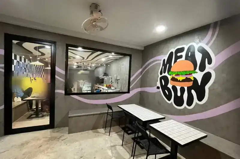 Inside the shop: grey walls with a lilac stripe, the Meat & Bun logo painted on the wall, white tiled tables, and the black-and-white swirl ceiling seen through the window