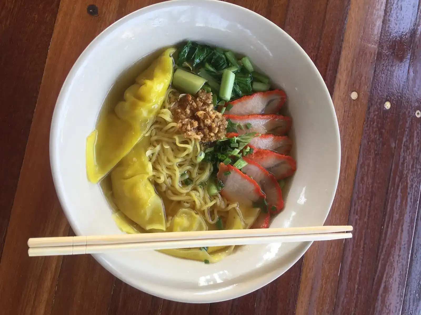 Egg noodles with wontons, red pork and greens in clear soup, chopsticks across the bowl