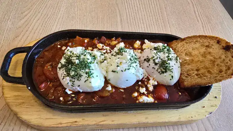 Shakshuka in a black cast-iron pan: three poached eggs in tomato stew with crumbled cheese, herbs and a slice of toast