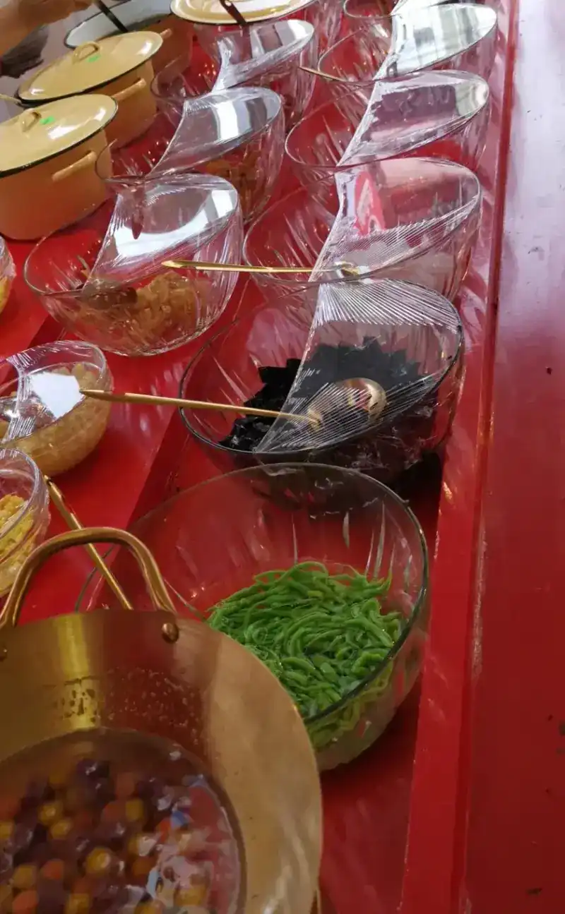 The topping counter: enamel pots, glass bowls of toppings, green lod chong and a brass bowl of coloured round sweets
