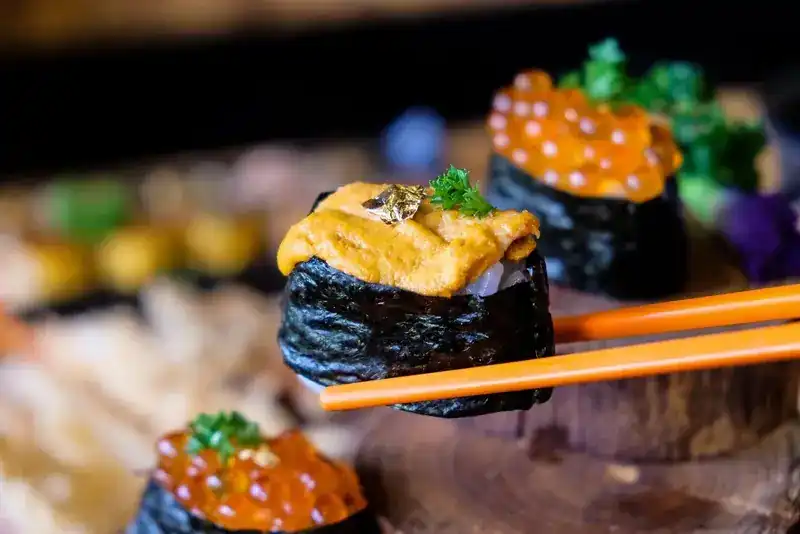 Sea urchin gunkan sushi held in orange chopsticks, salmon roe gunkan behind