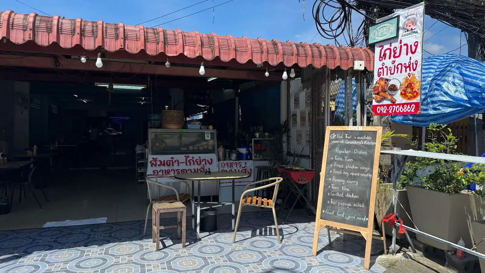 The shop front: a pink awning, tables on a blue tiled floor, a chalkboard of popular dishes and the red grilled chicken and papaya salad banner