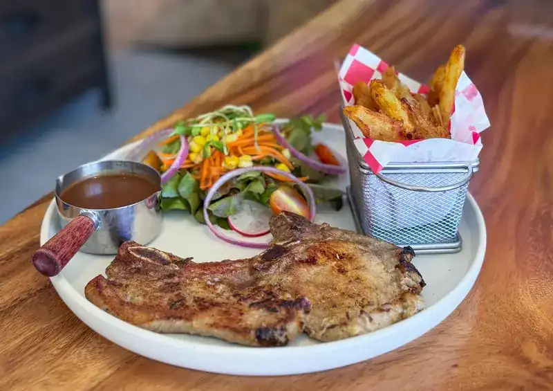 A grilled chop on a white plate with salad, a small pot of gravy and a basket of fries