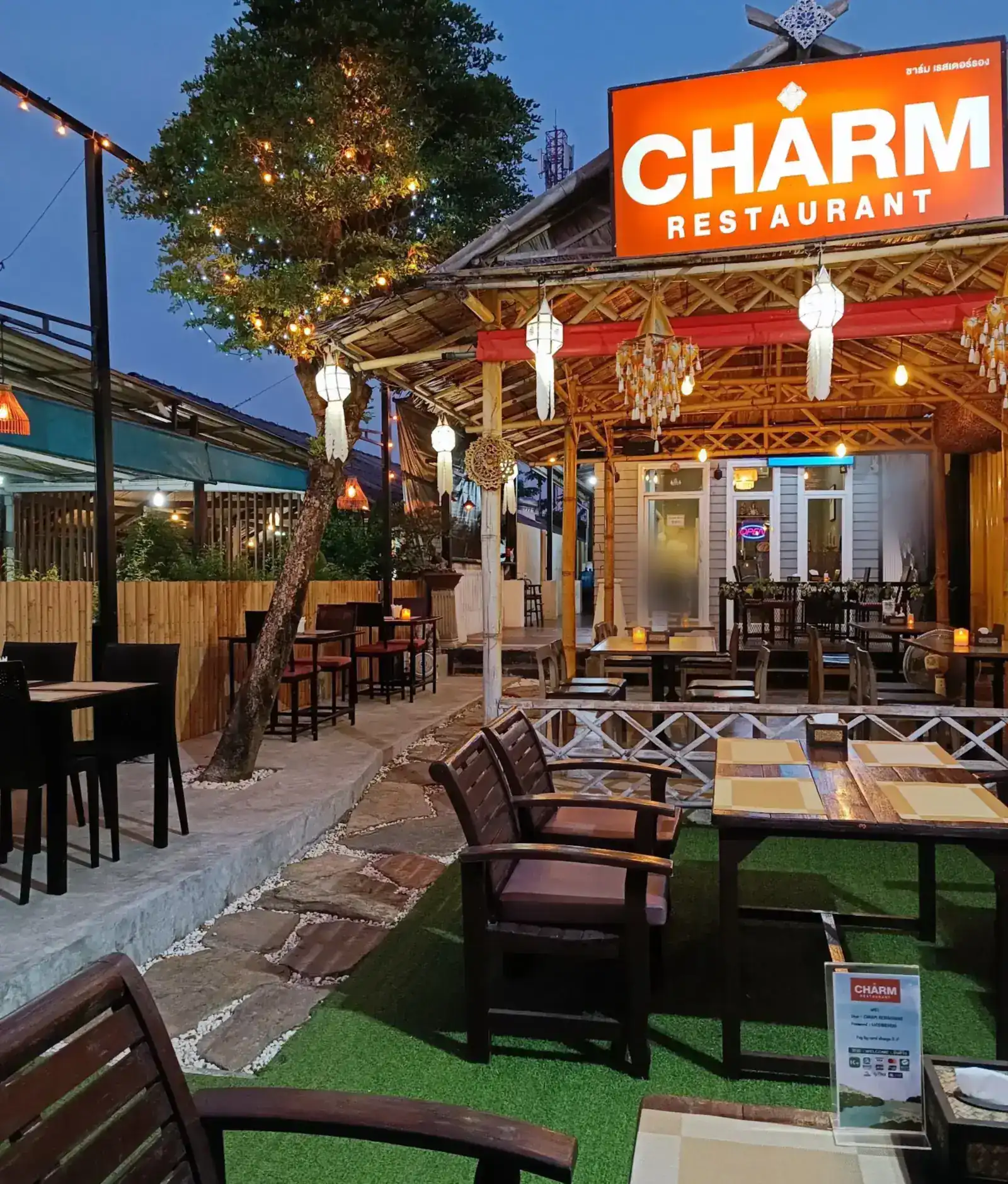 Charm Restaurant at dusk: the lit orange sign above a bamboo pavilion hung with lanterns, a tree strung with small lights, and wooden armchairs on a green lawn