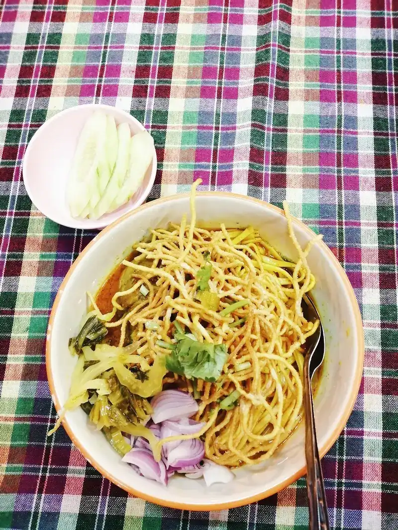 A bowl of khao soi seen from above, with pickled greens and red onion, on a checked cloth