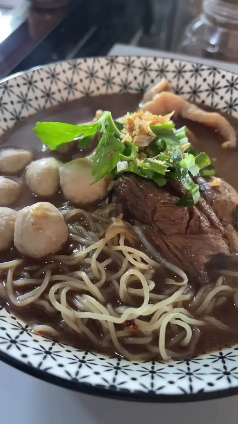 Egg noodles with braised beef, meatballs and herbs in a dark broth, in a blue and white patterned bowl