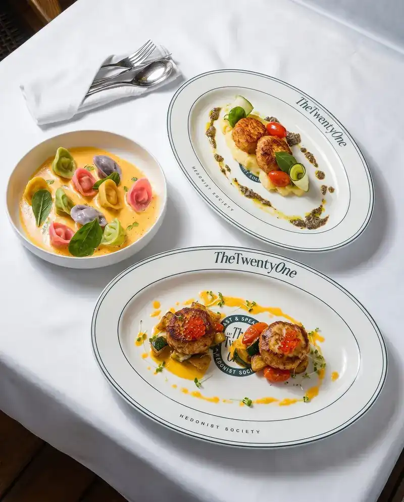 Three dishes on white linen: coloured tortellini in a golden sauce and two rimmed oval plates lettered TheTwentyOne