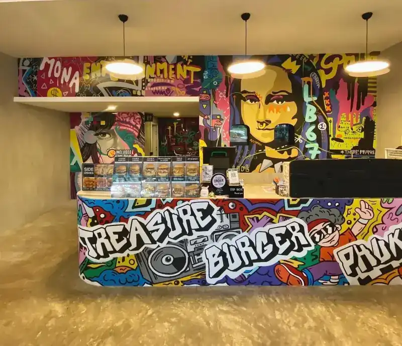 The counter painted in graffiti with Treasure Burger Phuket lettering, murals on the wall behind / เคาน์เตอร์ลายกราฟฟิตีพร้อมชื่อร้าน และภาพวาดบนผนังด้านหลัง