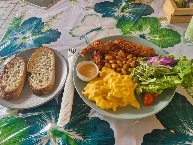 Scrambled eggs, baked beans, bacon, a sausage and salad with two slices of bread, on a flower-print tablecloth.