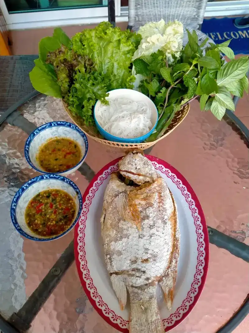 A whole fish with a basket of lettuce, herbs and rice noodles, and two bowls of dipping sauce