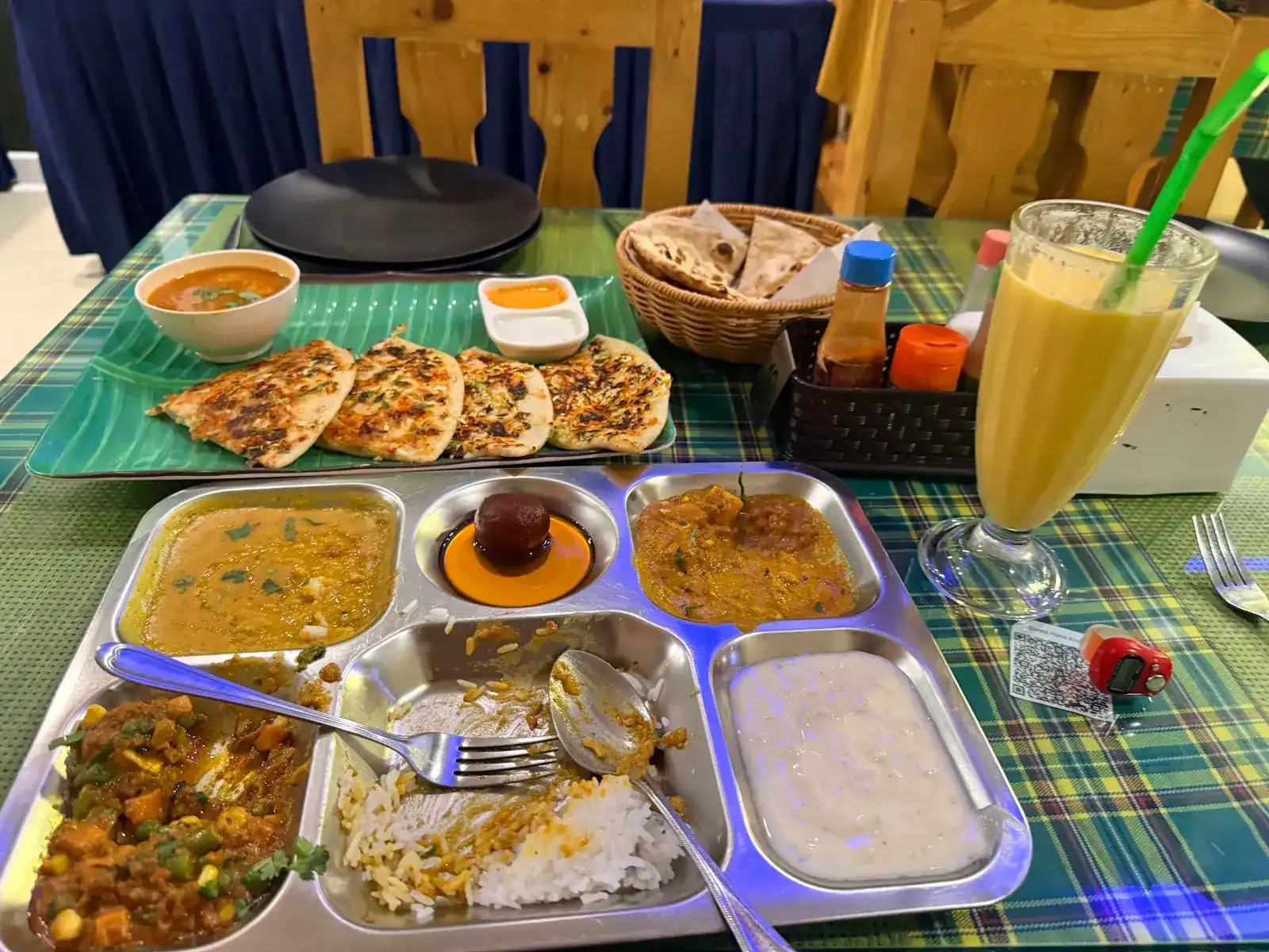 A steel thali, stuffed parathas on a leaf-green tray, a basket of roti and a lassi on a green check tablecloth