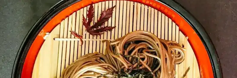 Soba noodles with shredded nori on a round bamboo tray with a red rim, garnished with a red maple leaf