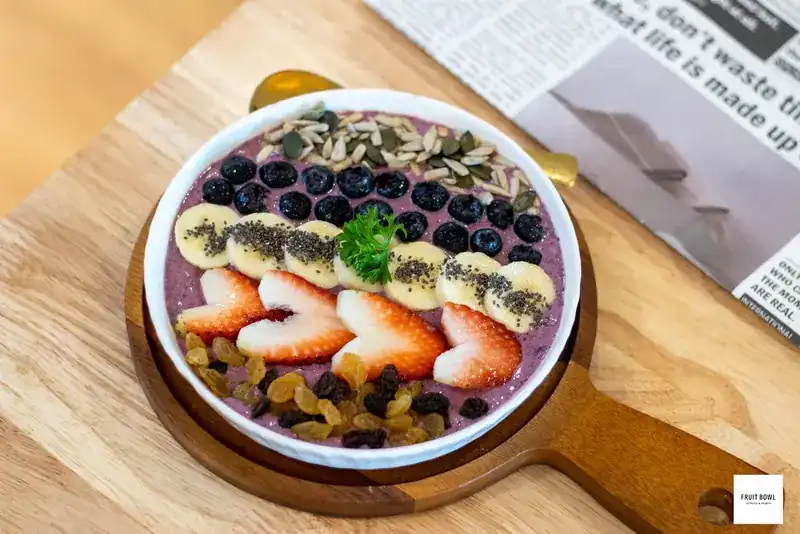 A smoothie bowl topped with blueberries, banana, heart-cut strawberries, seeds and raisins