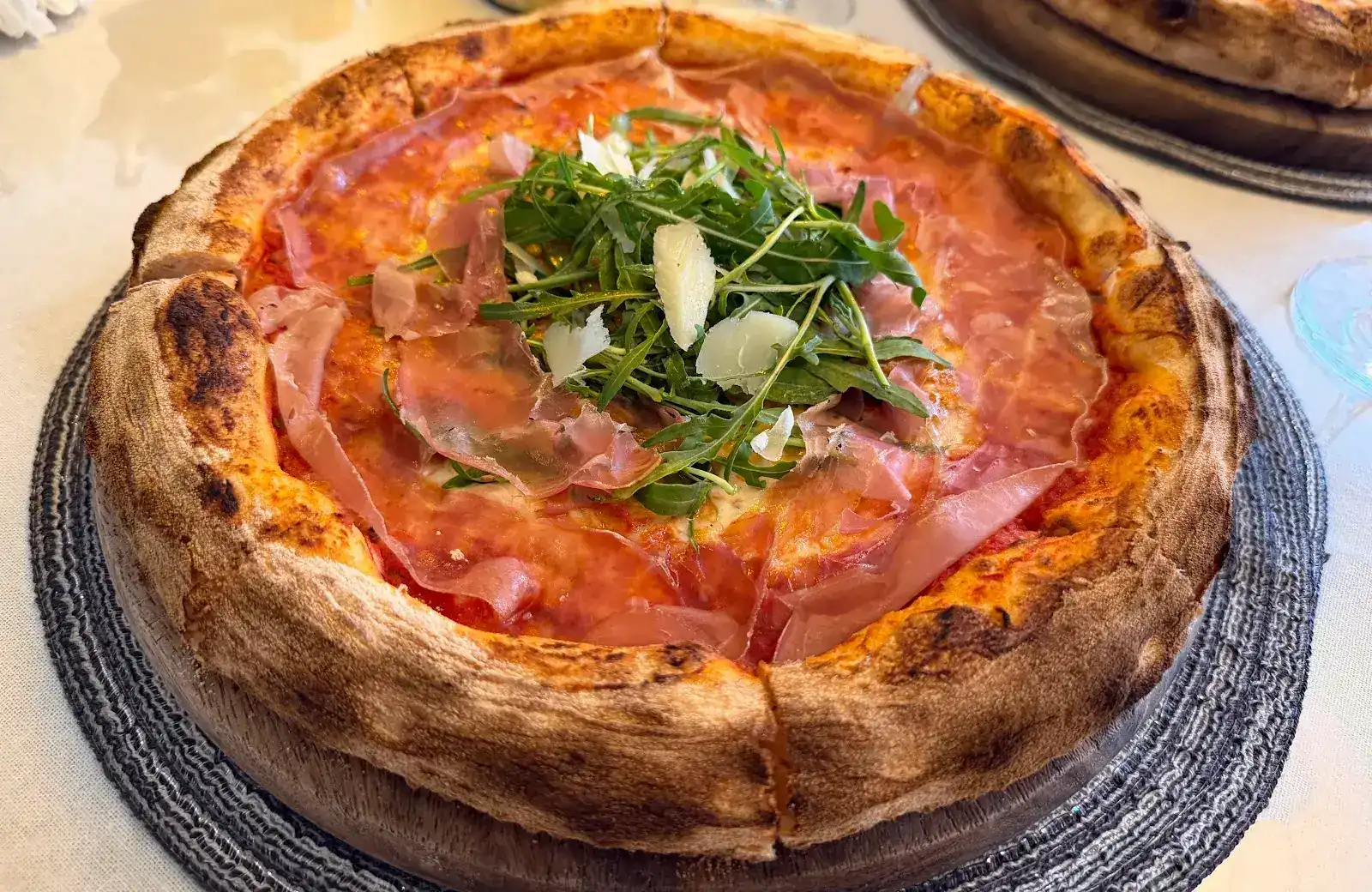 Neapolitan-style pizza with thin slices of cured ham, rocket and cheese shavings, its tall crust blistered by the oven