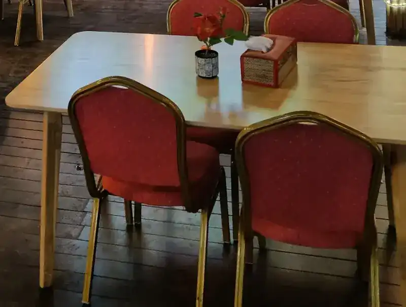 A table with a single red rose and a woven tissue box, red chairs with gold frames drawn up to it