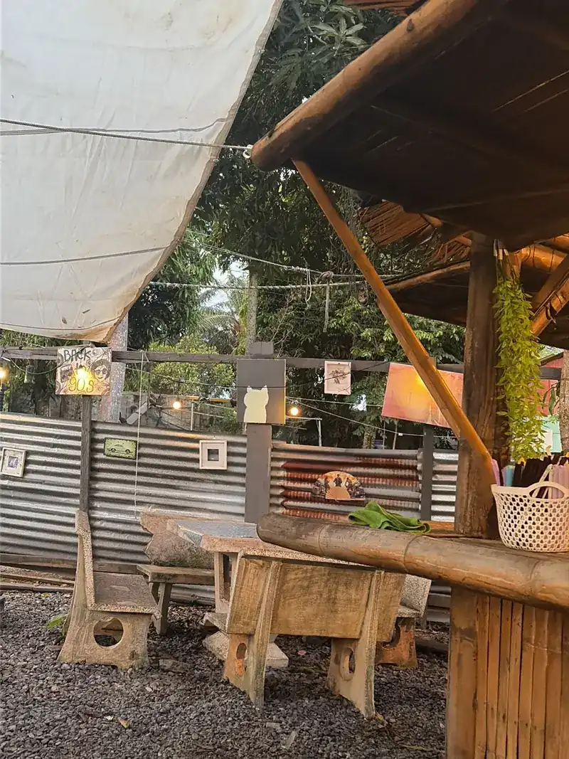 The garden by day: wooden benches on gravel, a bamboo bar and a canvas sail under the trees