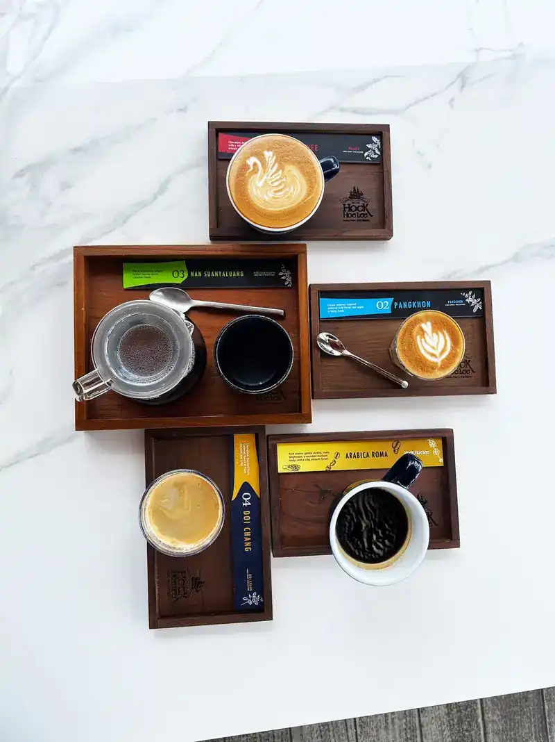 Five coffees on wooden trays seen from above, each beside a card naming its bean: Nan Suanyaluang, Pangkhon, Doi Chang and Arabica Roma