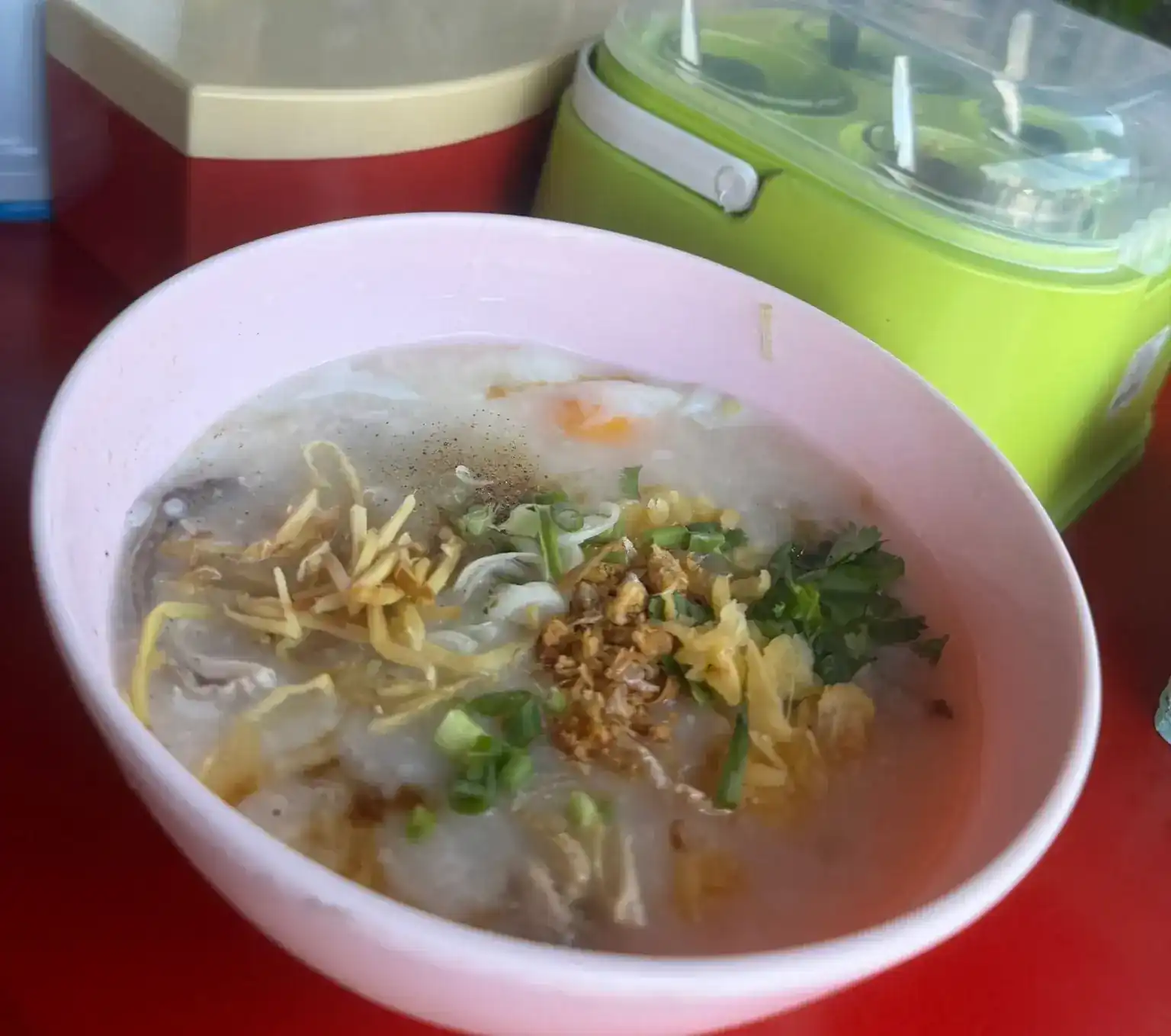 A pink bowl of rice porridge with a soft egg, ginger, spring onion, coriander and fried garlic on a red table