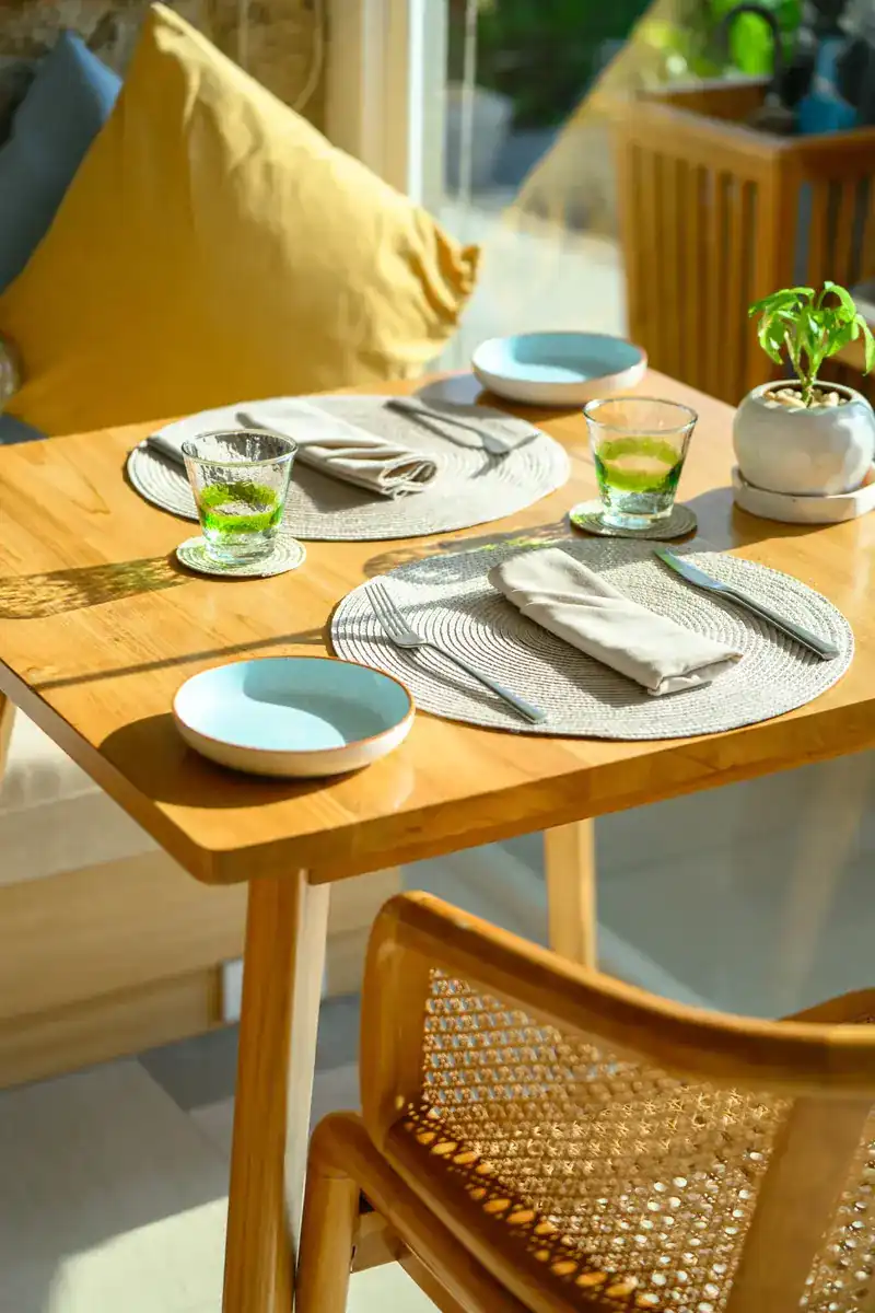 A sunlit wooden table laid for two with blue plates, woven mats and a yellow cushion