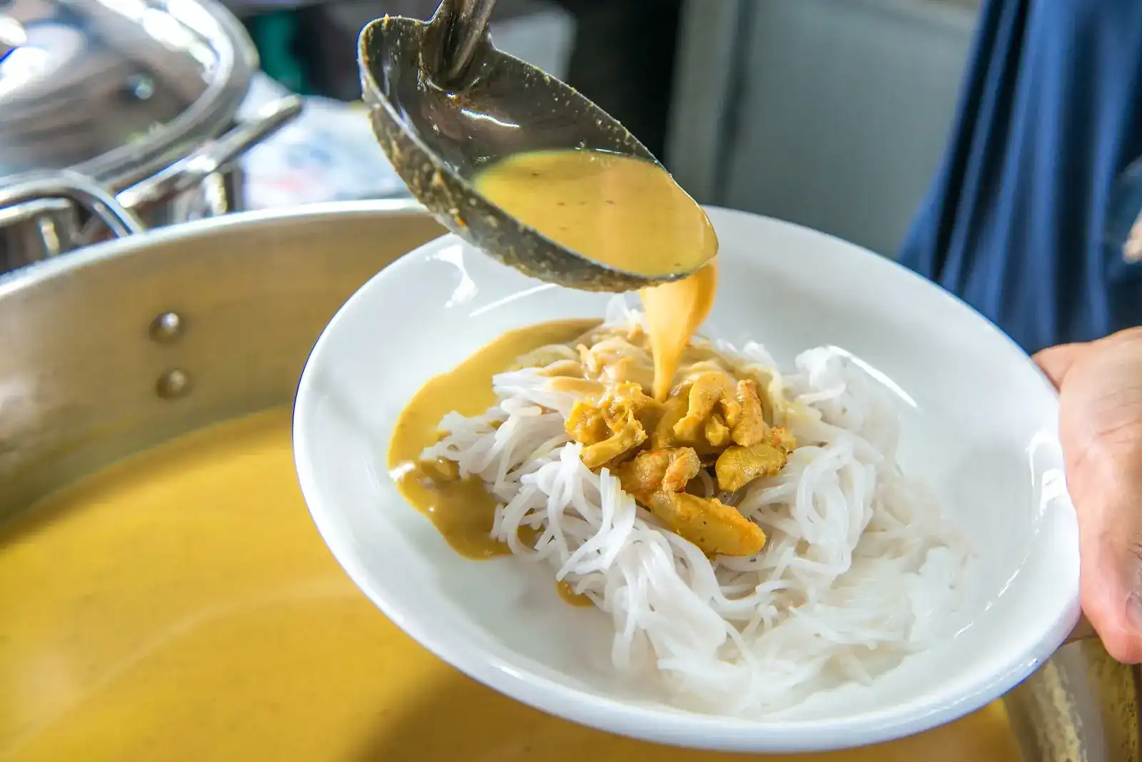 A ladle of yellow curry poured over a plate of fresh khanom jeen noodles