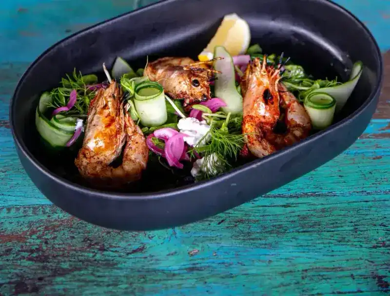 Grilled prawns with cucumber ribbons and dill in a black bowl on a painted teal table