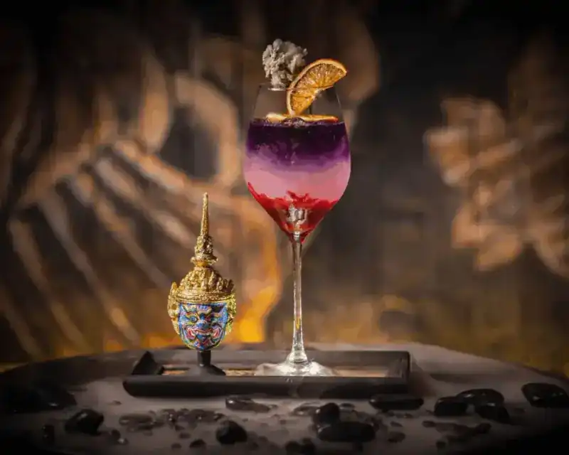 A layered purple and pink cocktail beside a miniature Thai khon mask