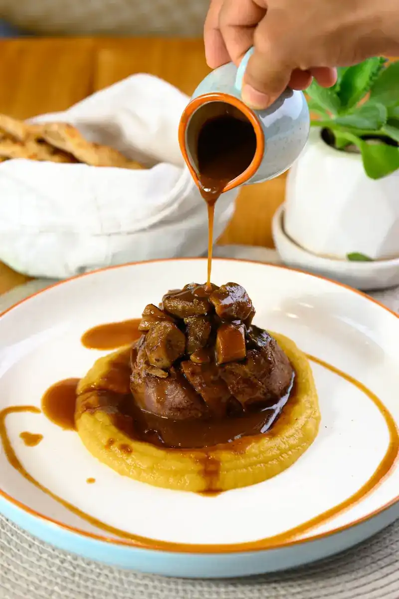 Sauce poured from a small jug over meat on a bed of purée