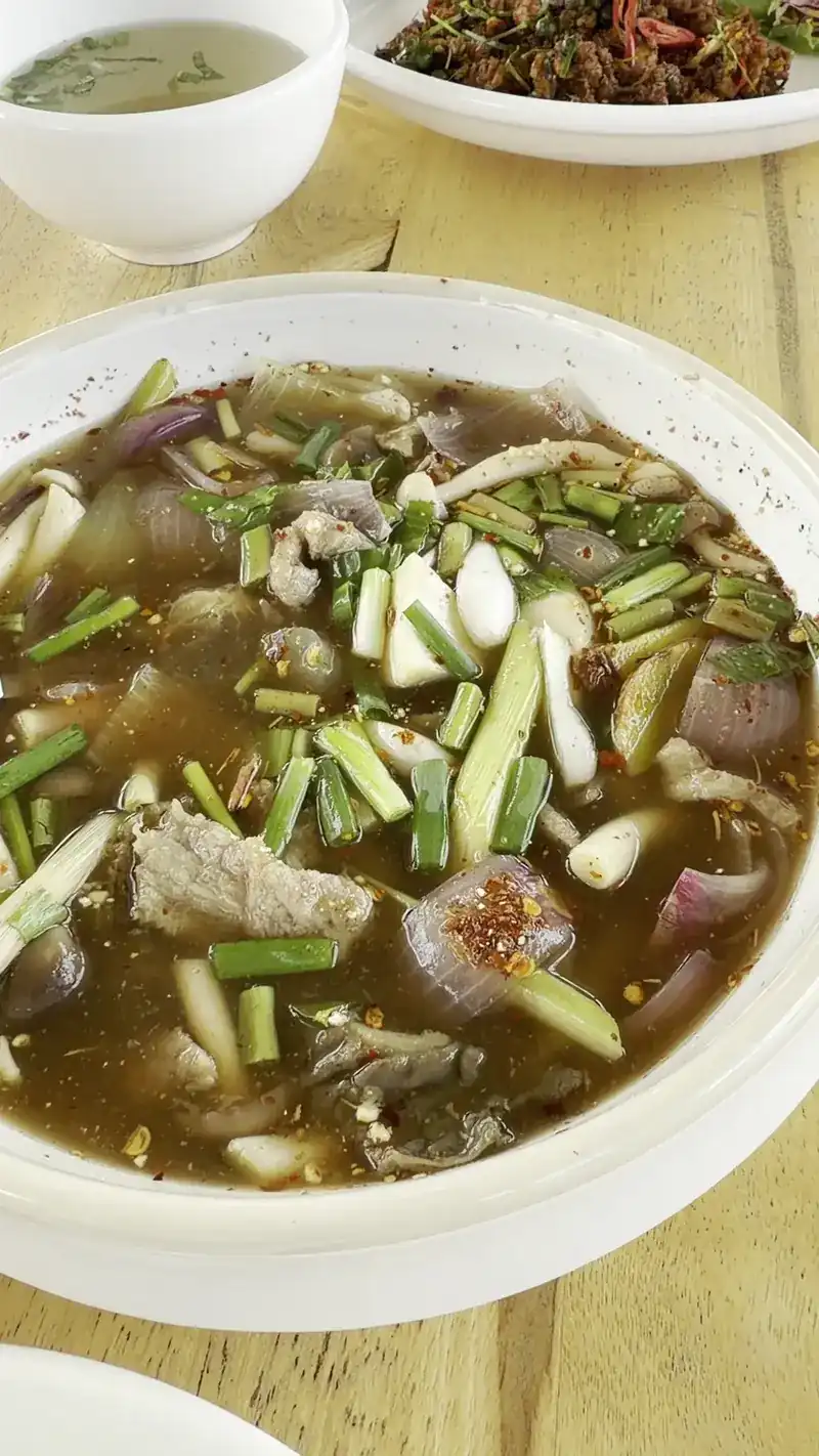 A wide bowl of clear hot and sour soup with meat, lemongrass and spring onion on a wooden table