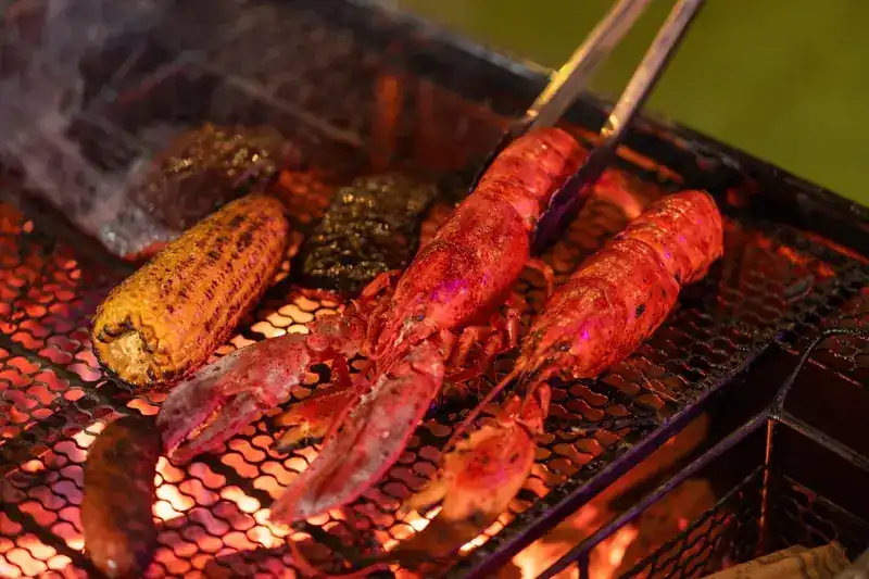 Two lobsters, corn and sausages on a mesh grill over glowing coals