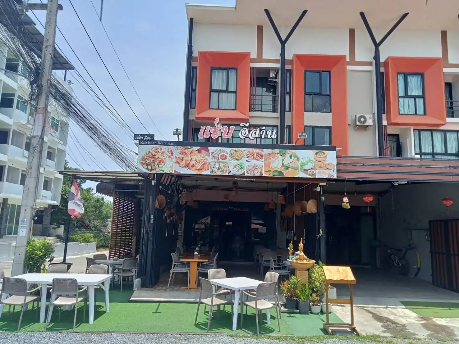 The front of Zab Isaan: a shophouse with orange window frames, the pink แซ่บ อีสาน sign, woven baskets under the awning and white tables out front on green turf