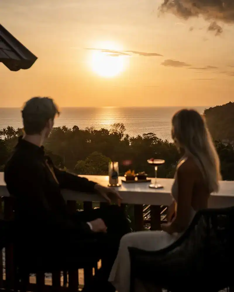 Two guests seen from behind at the terrace rail, the sun low over the Andaman Sea