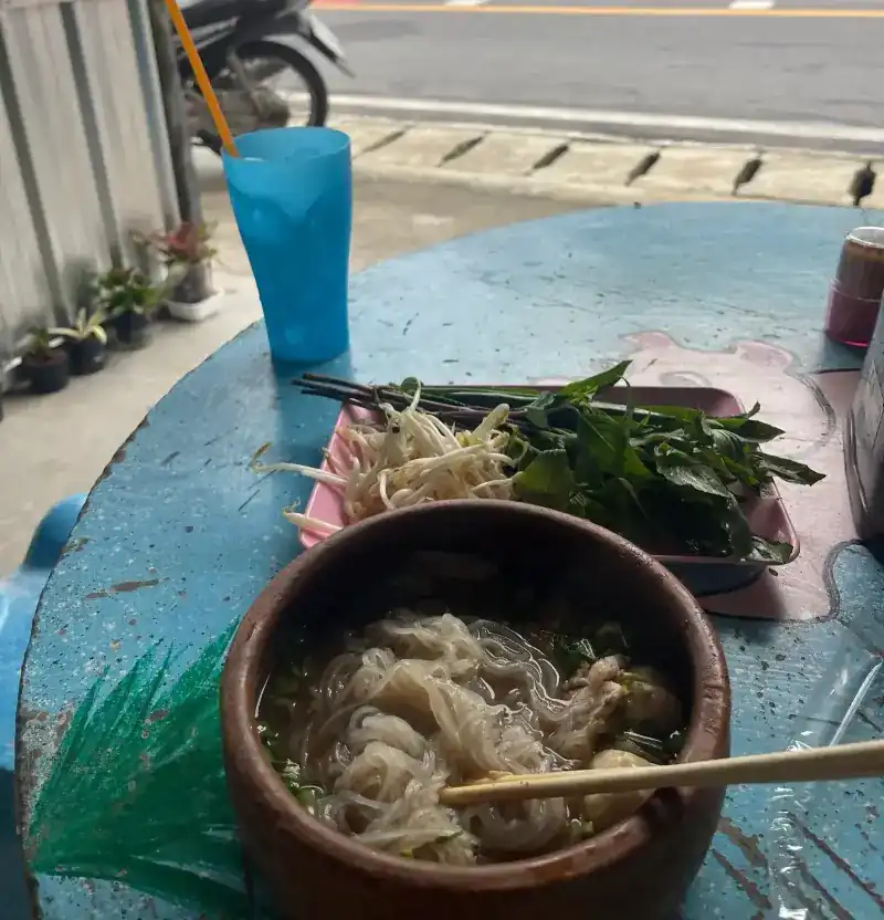 A clay pot of noodles on a blue table by the road, with bean sprouts, herbs and a blue cup