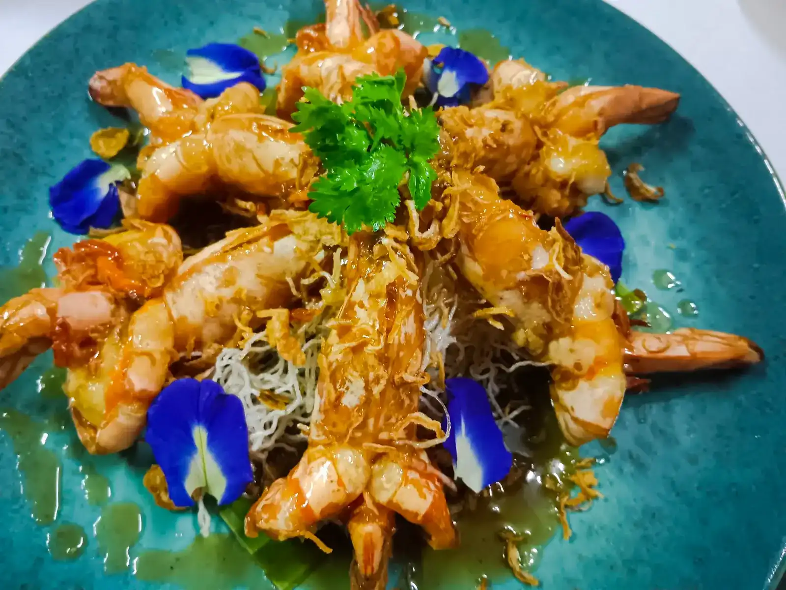 Prawns on crispy fried noodles with butterfly-pea flowers, on a blue-green plate