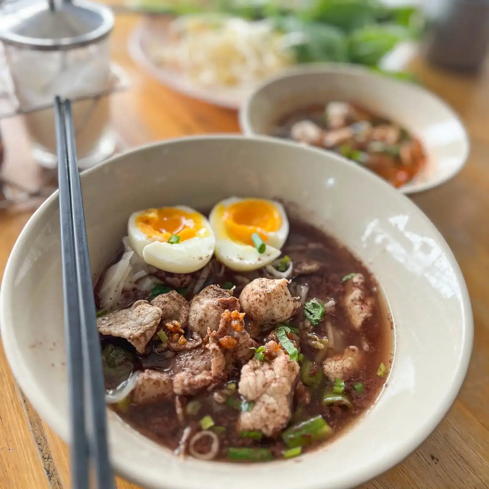 Boat noodle soup in a dark broth with pork, meatballs and two soft-boiled eggs, chopsticks resting on the bowl