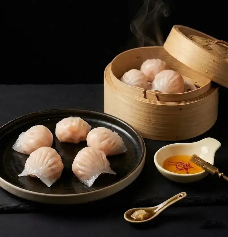 Ha gow shrimp dumplings on a dark plate beside a bamboo steamer