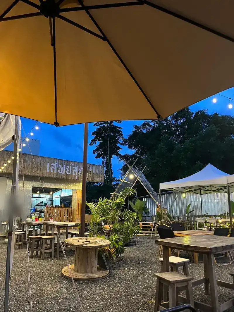 Under a big umbrella: the kitchen with its เสพย์สุก sign, wooden tables and a white canopy