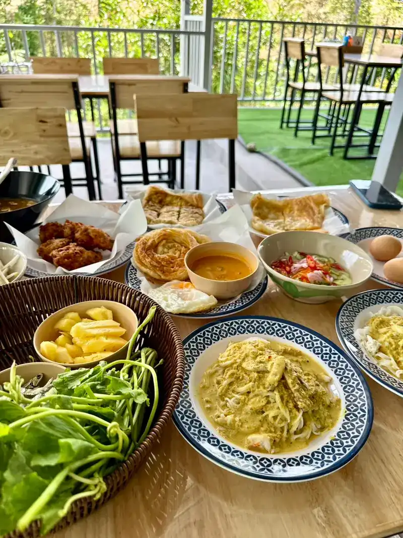 A full table by the terrace: khanom chin with curry, a basket of fresh greens, roti, fried chicken and pineapple