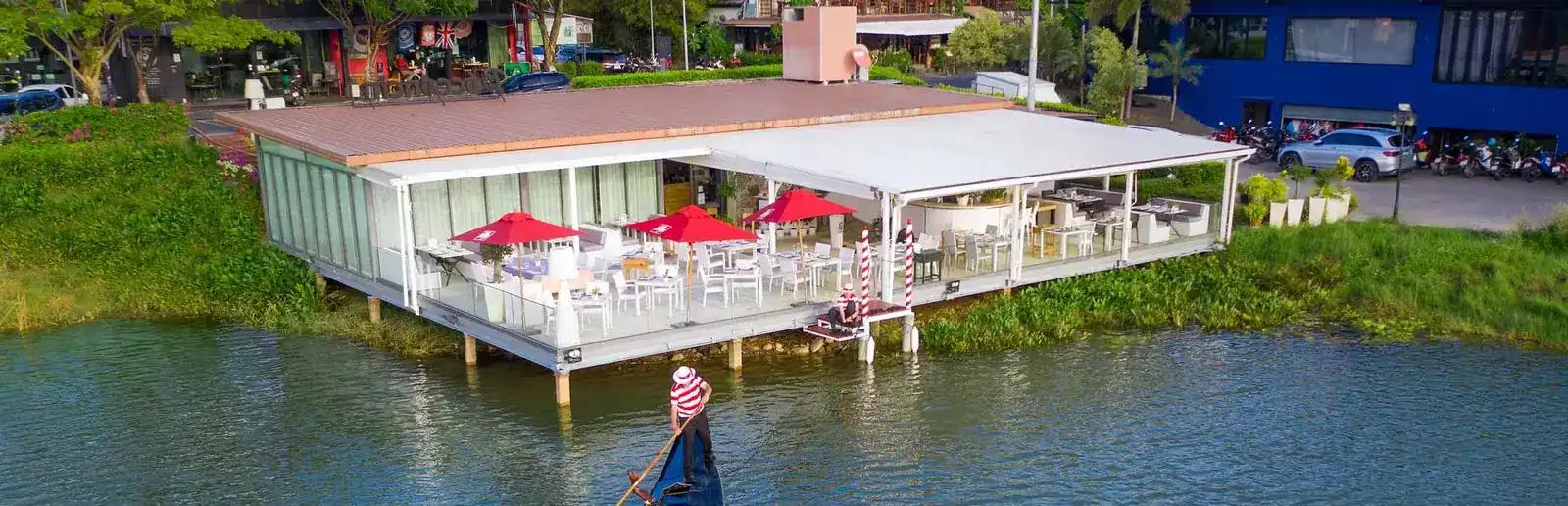 The white terrace of Bocconcino standing on stilts over the lake, red parasols open, a gondolier in a striped shirt rowing past