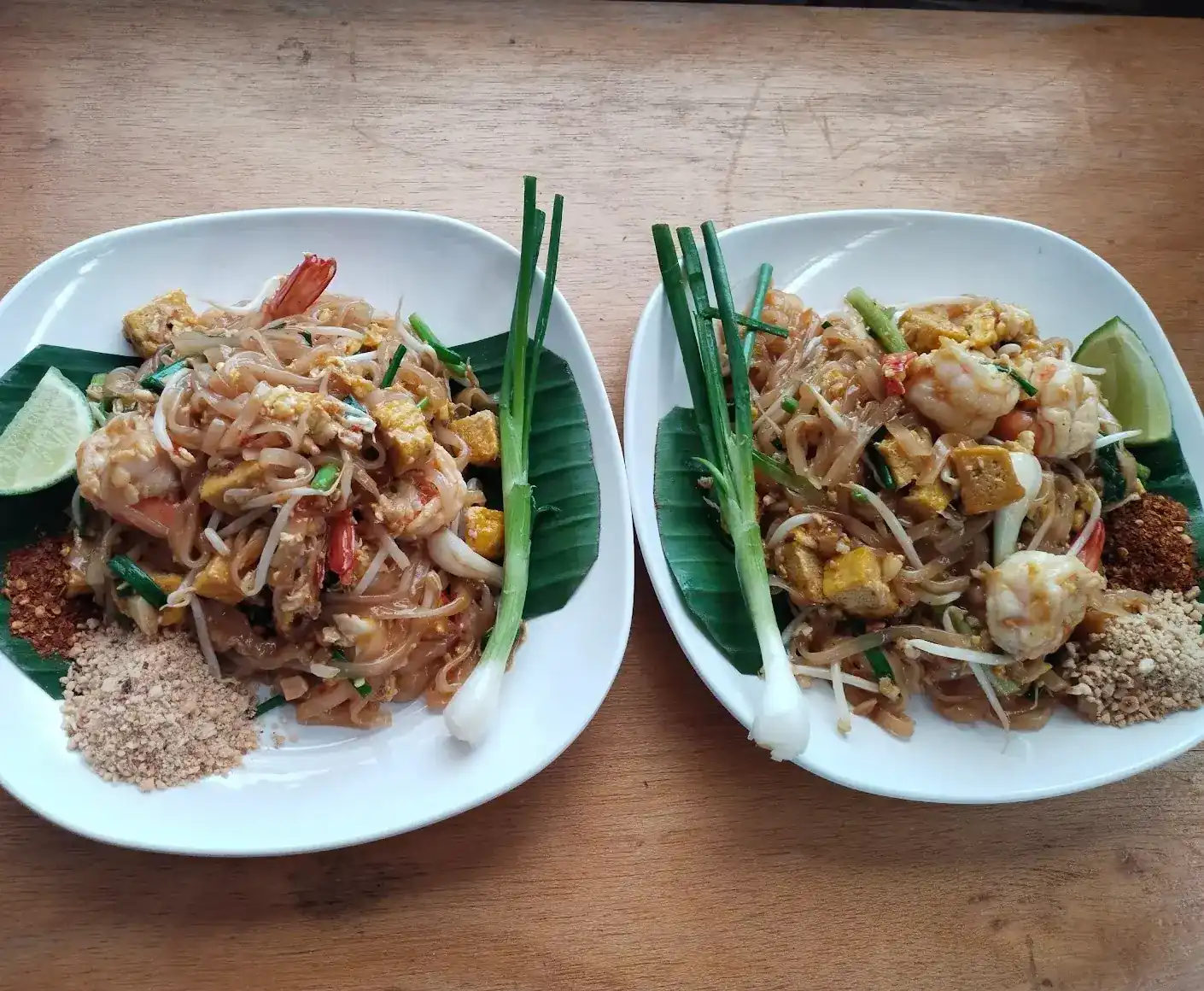Two plates of pad thai with prawns on banana leaf, with lime, ground peanuts and chilli flakes on the side