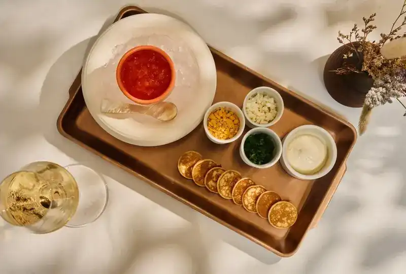A tray of blini with red caviar and condiments beside a glass of white wine